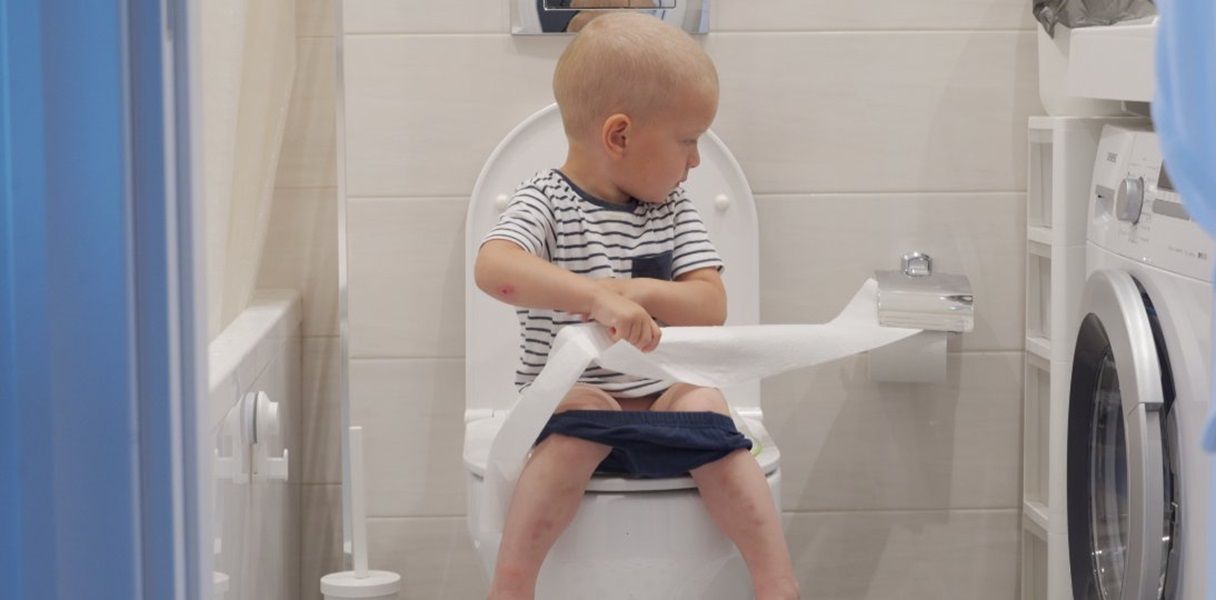 A boy sitting on the toilet and unrolling toilet paper