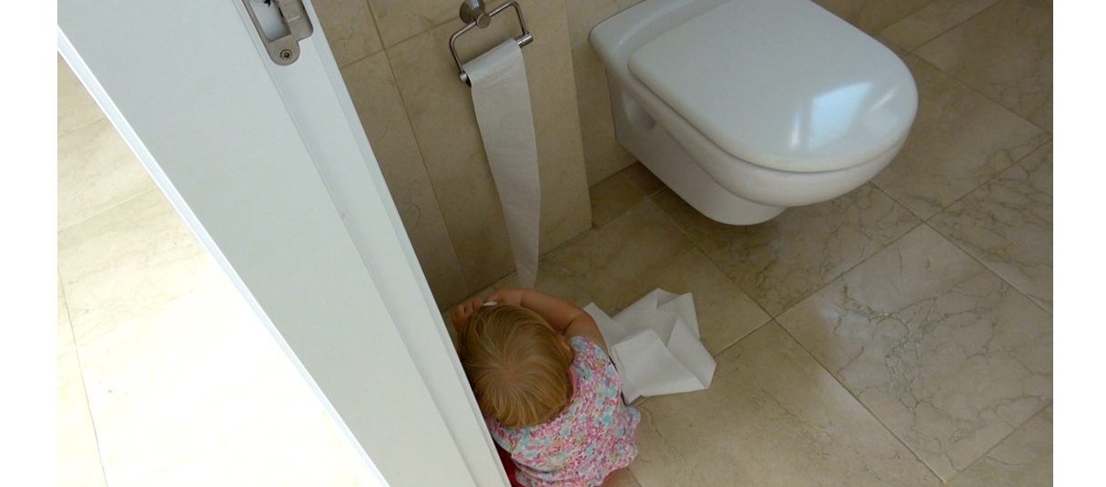 A toddler sitting next to the toilet roling down toilet paper
