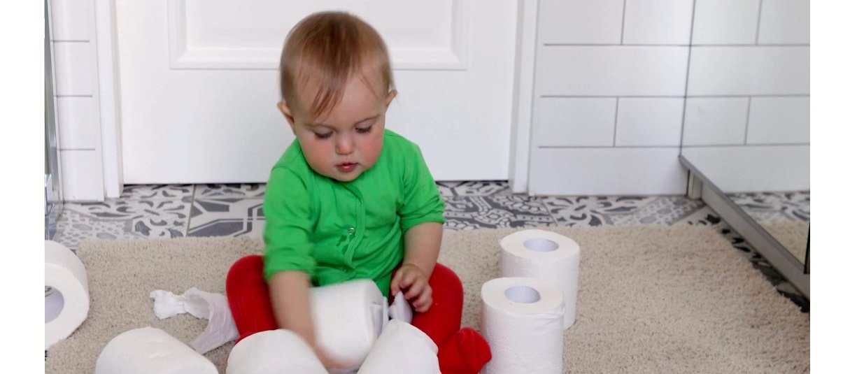 A toddler sitting in a bathroom playing with toilet paper