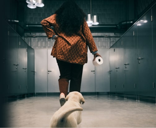 Woman holding a toilet roll walking into ladies room followed by a labrador puppy