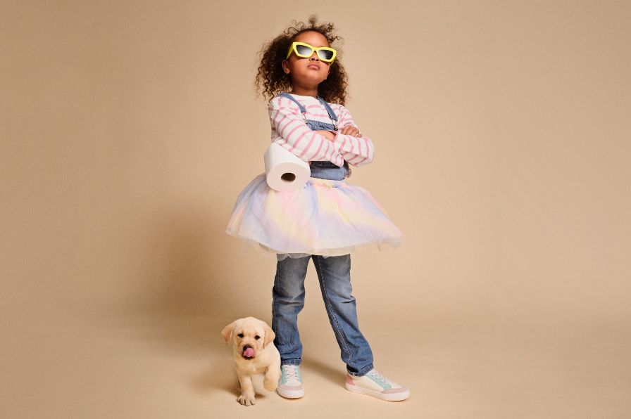A confident girl wearing sunglasses, standing beside a puppy while holding a toilet roll.